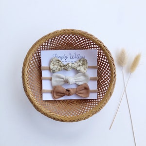Puede incluir: Conjunto de tres lazos para el pelo en una cesta de mimbre. Los lazos están hechos de tela con estampado floral, beige liso y marrón liso. Los lazos están anudados en el centro. El texto "Simply Willow Designs" está impreso en una tarjeta blanca detrás de los lazos.