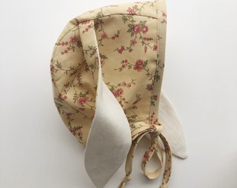 May include: A yellow bonnet with a floral print featuring red roses and green leaves. The bonnet has a white lining and ties at the back.
