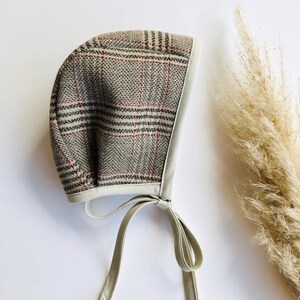 May include: A plaid baby bonnet with a white trim and matching ties. The bonnet is made of a soft, woven fabric and has a classic, vintage style.