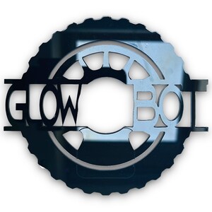 Może przedstawiać: Czarny, okrągły metalowy znak z wyciętym tekstem "GLOW BOI". Znak ma zębatą zewnętrzną krawędź i okrągły wewnętrzny wzór. Litery są pogrubione i bezszeryfowe.