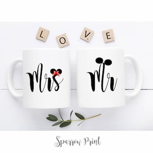 May include: Two white ceramic mugs with black lettering. The mug on the left says "Mrs." with a red bow and the mug on the right says "Mr." with two black Mickey Mouse ears.