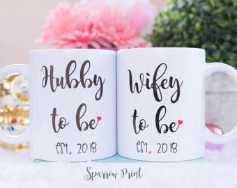 May include: Two white ceramic mugs with black text that reads "Hubby to be est. 2018" and "Wifey to be est. 2018".  Each mug has a small red heart next to the text.