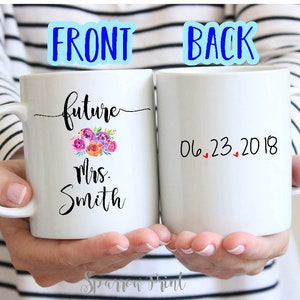 May include: Two white ceramic mugs with black text. The front mug says "future Mrs. Smith" with a floral design. The back mug says "06.23.2018" with three red hearts.