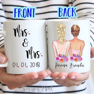 May include: Two white ceramic mugs with black text and illustrations. The front mug says "Mrs. & Mrs. 01.01.2018". The back mug has an illustration of two women in wedding dresses with flowers and the text "Karina Brenda".