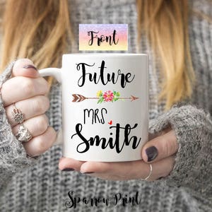 K&ouml;nnte beinhalten: Eine wei&szlig;e Keramiktasse mit einem schwarzen Pfeil und einem Blumenmuster. Der Text "Future Mrs. Smith" ist auf die Tasse gedruckt.