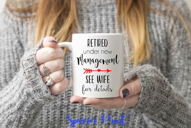 May include: White ceramic mug with black text that reads "Retired under new management see wife for details". A red arrow with a heart points to the right. The mug is being held by a person wearing a gray sweater.
