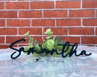 May include: Black wooden sign with the name Samantha written in a cursive font. The sign is sitting on a white surface with a brick wall in the background.