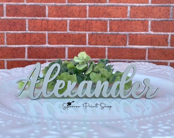 May include: White wooden name sign with the name "Alexander" written in cursive script. The sign is on a white plate with green foliage behind it.