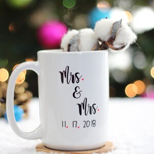 May include: White ceramic mug with black text that reads "Mrs. & Mrs. 11.17.2018" with small red hearts between the numbers. The mug is on a wooden coaster and a white fuzzy surface.
