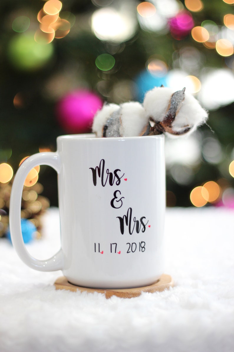 May include: White ceramic mug with black text that reads "Mrs. & Mrs. 11.17.2018" with small red hearts between the numbers. The mug is on a wooden coaster and a white fuzzy surface.