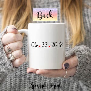 K&ouml;nnte beinhalten: Eine wei&szlig;e Keramiktasse mit schwarzem Text, der "06.22.2018" lautet, mit drei roten Herzen zwischen den Zahlen. Die Tasse wird von einer Person gehalten, die einen grauen Pullover tr&auml;gt.