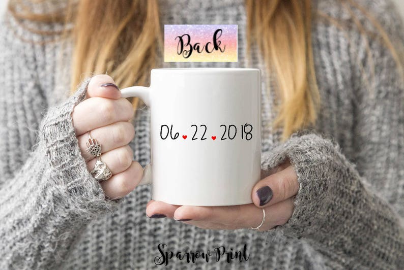 K&ouml;nnte beinhalten: Eine wei&szlig;e Keramiktasse mit schwarzem Text, der "06.22.2018" lautet, mit drei roten Herzen zwischen den Zahlen. Die Tasse wird von einer Person gehalten, die einen grauen Pullover tr&auml;gt.