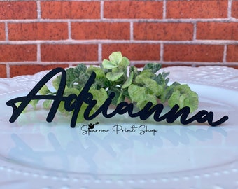 May include: Black wooden name card with the name "Arianne" written in a cursive script. The card is on a white plate with green foliage behind it. The text "Sparrow Print Shop" is visible below the name.