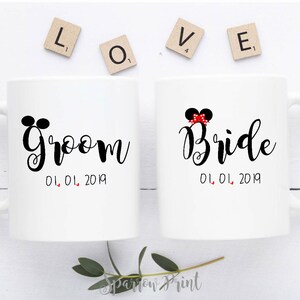 May include: Two white ceramic mugs with black handles. The mug on the left has the word "Groom" written in black cursive with Mickey Mouse ears above the word. The mug on the right has the word "Bride" written in black cursive with a red bow and Mickey Mouse ears above the word. Both mugs have the date "01.01.2019" written below the word.