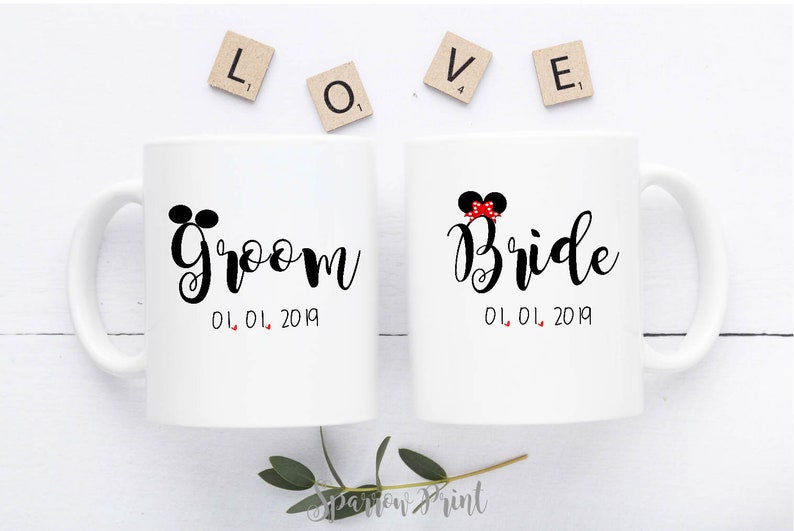 May include: Two white ceramic mugs with black handles. The mug on the left has the word "Groom" written in black cursive with Mickey Mouse ears above the word. The mug on the right has the word "Bride" written in black cursive with a red bow and Mickey Mouse ears above the word. Both mugs have the date "01.01.2019" written below the word.