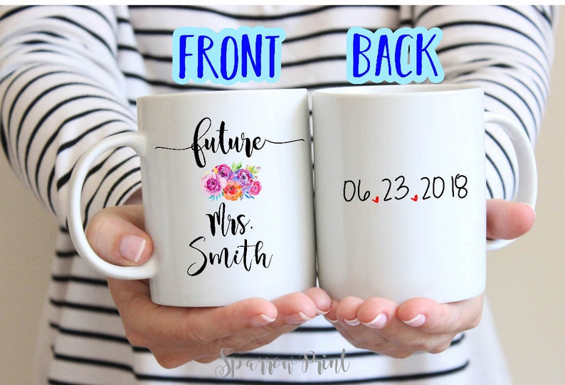 May include: Two white ceramic mugs with black text. The front mug says "future Mrs. Smith" with a floral design. The back mug says "06.23.2018" with three red hearts.