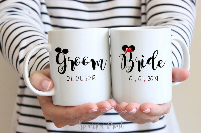 May include: Two white ceramic mugs with black text and red accents. The mug on the left says "Groom 01.01.2019" and the mug on the right says "Bride 01.01.2019".