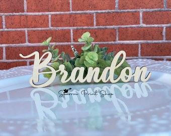 May include: Wooden name place card with the name "Brandon" written in cursive script. The place card is light brown and is sitting on a white surface.