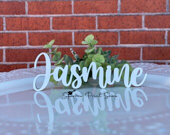 May include: White wooden name sign with the word "Jasmine" written in a cursive font. The sign is sitting on a white surface with a brick wall in the background.