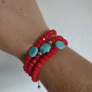 Puede incluir: Una pulsera de cuentas rojas con tres hilos, adornada con piedras ovaladas de color turquesa y pequeñas cuentas doradas. La pulsera está enrollada alrededor de una muñeca, mostrando su diseño y contraste de colores.