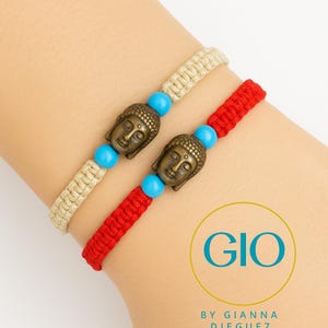 Puede incluir: Dos pulseras de macramé con abalorios de cabeza de Buda de bronce y cuentas turquesas. Una pulsera es beige, la otra es roja. Las pulseras se muestran en la muñeca de una persona. La imagen incluye el texto "GIO BY GIANNA DIEGUEZ."