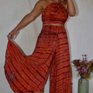 May include: An orange tie-dye halter top and wide-leg pants set. The fabric has a striped pattern with small, scattered embellishments. The pants have floral embroidery at the hem. The outfit is paired with platform boots.