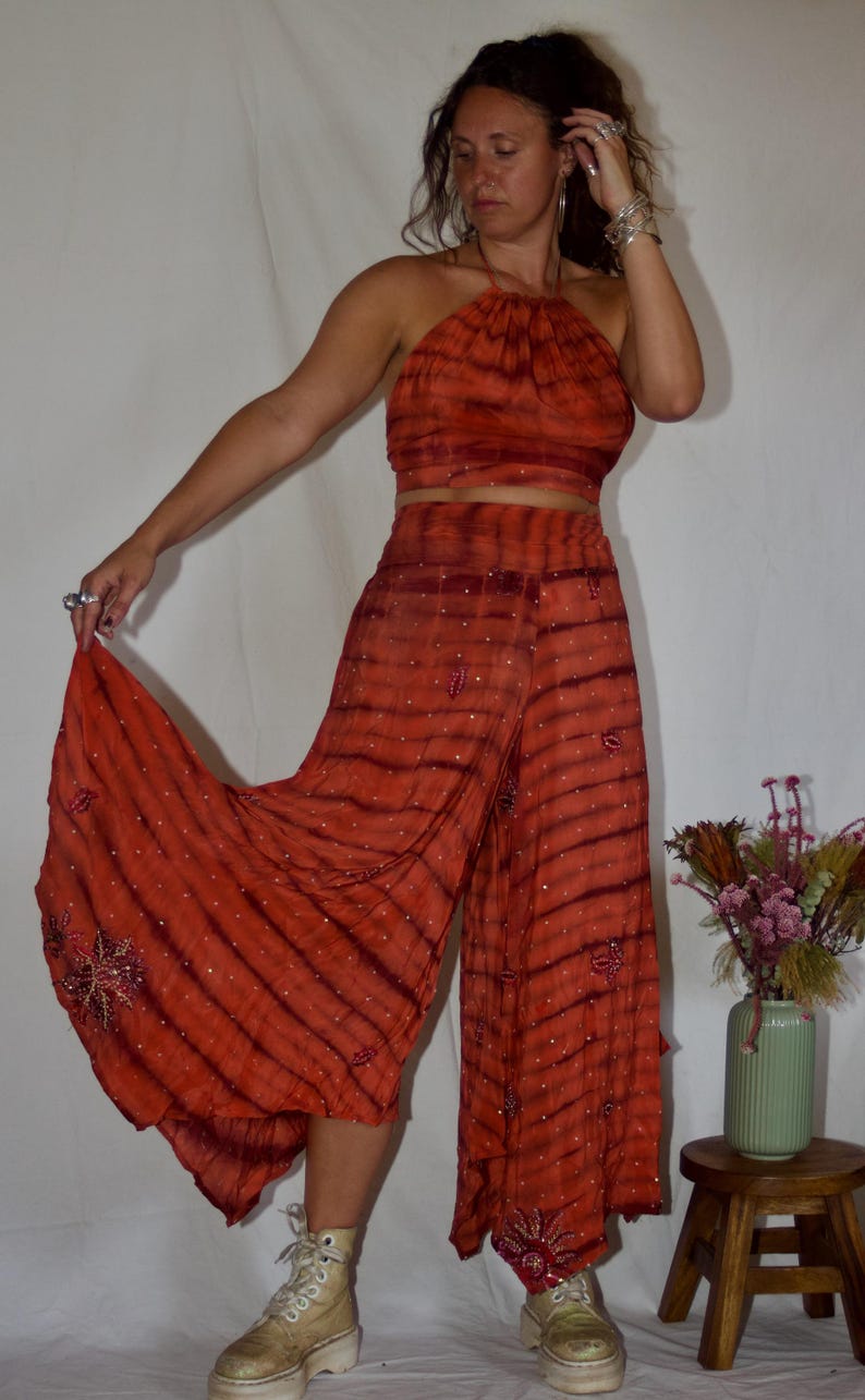 May include: An orange tie-dye halter top and wide-leg pants set. The fabric has a striped pattern with small, scattered embellishments. The pants have floral embroidery at the hem. The outfit is paired with platform boots.