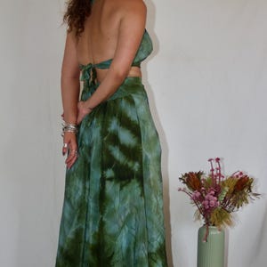 May include: A two-piece tie-dye outfit in shades of green and blue. The set includes a halter top and a flowing skirt. The outfit is paired with platform boots. A vase of dried flowers sits on a small wooden stool.