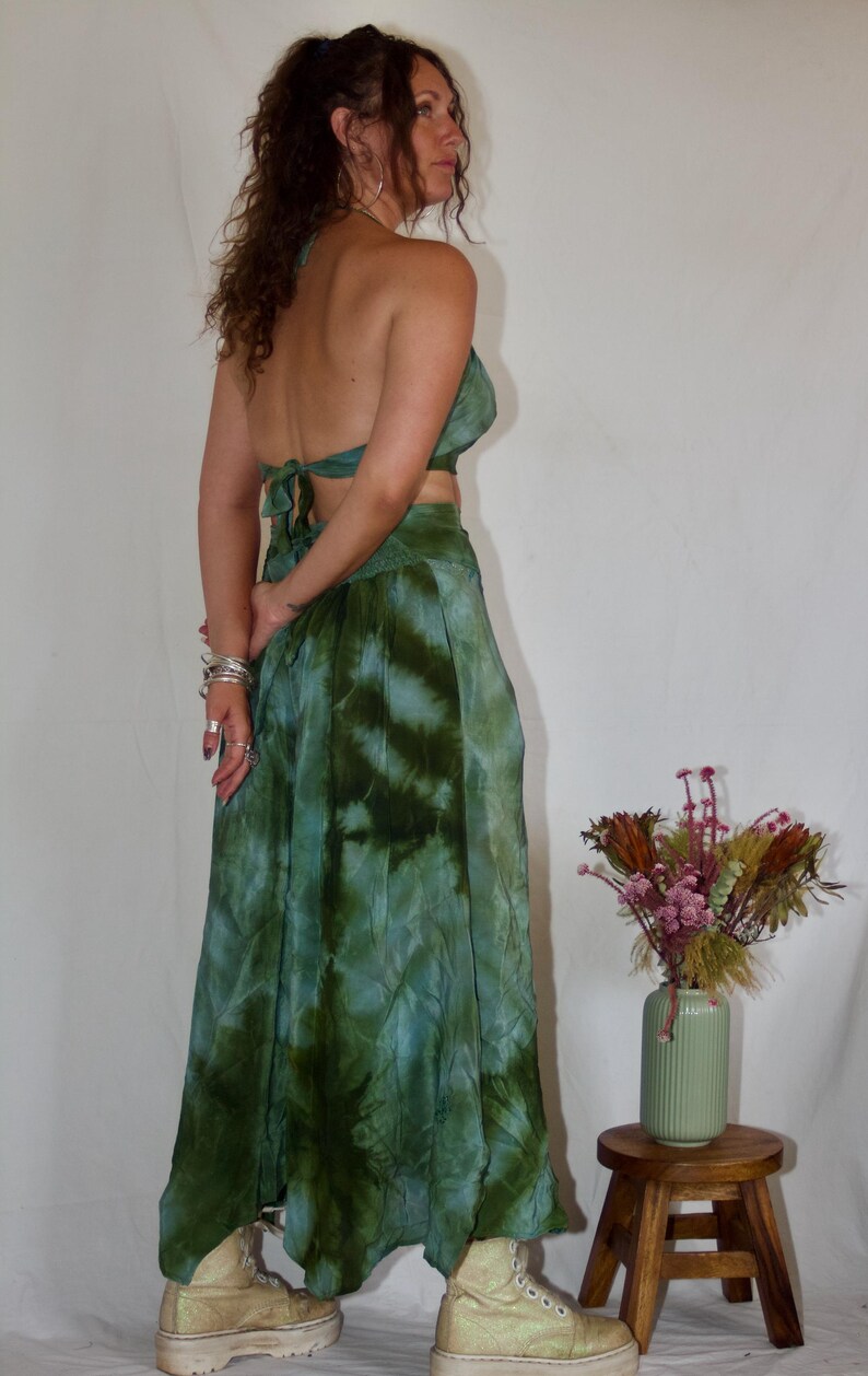 May include: A two-piece tie-dye outfit in shades of green and blue. The set includes a halter top and a flowing skirt. The outfit is paired with platform boots. A vase of dried flowers sits on a small wooden stool.