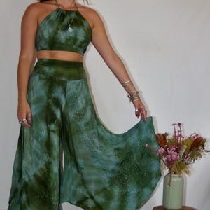 May include: A two-piece tie-dye outfit in shades of green and blue. The halter top and wide-leg pants have a flowing, bohemian style. The outfit is paired with platform boots. A small wooden stool and vase of flowers are in the background.
