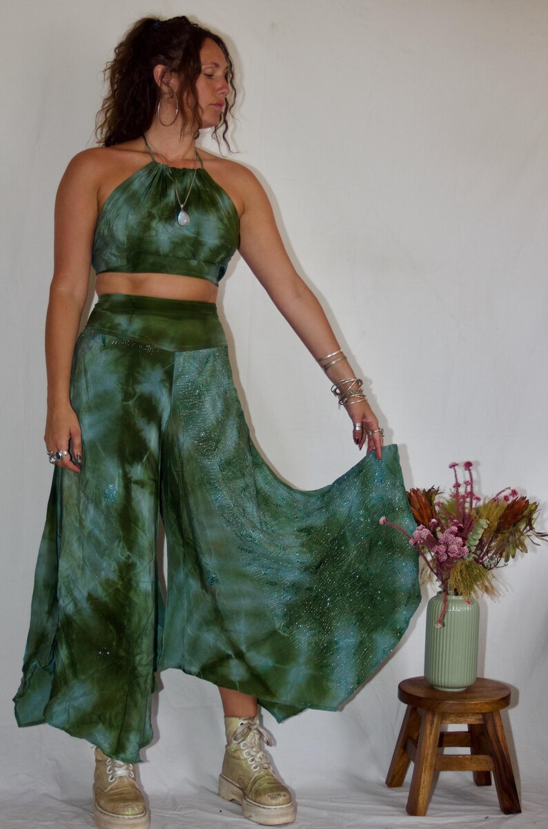May include: A two-piece tie-dye outfit in shades of green and blue. The halter top and wide-leg pants have a flowing, bohemian style. The outfit is paired with platform boots. A small wooden stool and vase of flowers are in the background.
