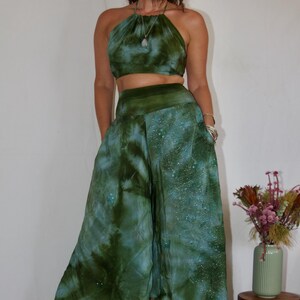 May include: A two-piece tie-dye outfit in shades of green and blue. The halter top and wide-leg pants feature a swirling pattern. The outfit is paired with platform boots. A small wooden stool holds a vase of dried flowers.