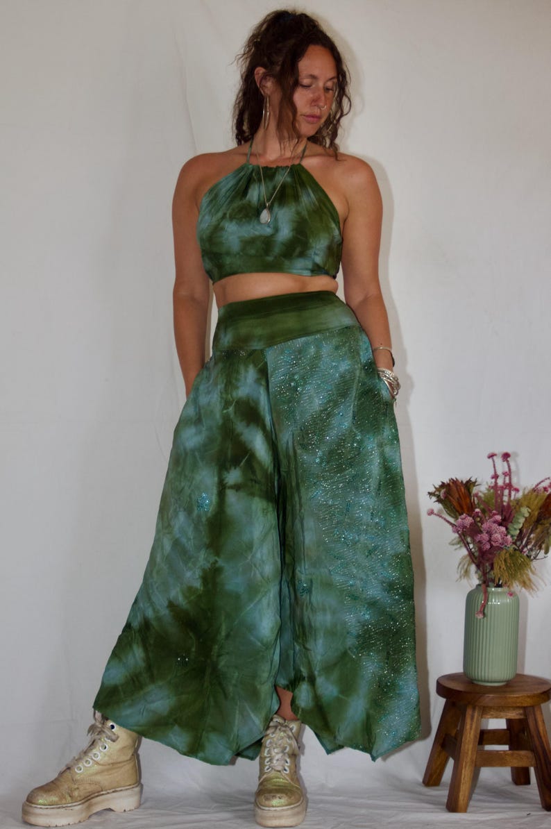 May include: A two-piece tie-dye outfit in shades of green and blue. The halter top and wide-leg pants feature a swirling pattern. The outfit is paired with platform boots. A small wooden stool holds a vase of dried flowers.