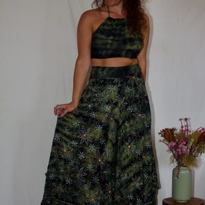 May include: A two-piece green tie-dye outfit. The halter top and long skirt are adorned with a floral pattern. The skirt has a wide hem with decorative beading. The model is wearing platform boots.