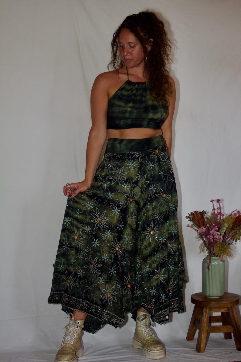 May include: A two-piece green tie-dye outfit. The halter top and long skirt are adorned with a floral pattern. The skirt has a wide hem with decorative beading. The model is wearing platform boots.