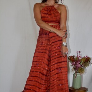 May include: A two-piece orange halter top and wide-leg pants set with a striped pattern and embellished details. The outfit is paired with platform boots. The ensemble is a bohemian style.