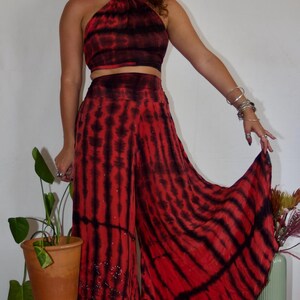 May include: A two-piece tie-dye outfit in red and black. The halter top and wide-leg pants feature a striped pattern. The pants have a beaded embellishment near the hem. The outfit is paired with light-colored boots.