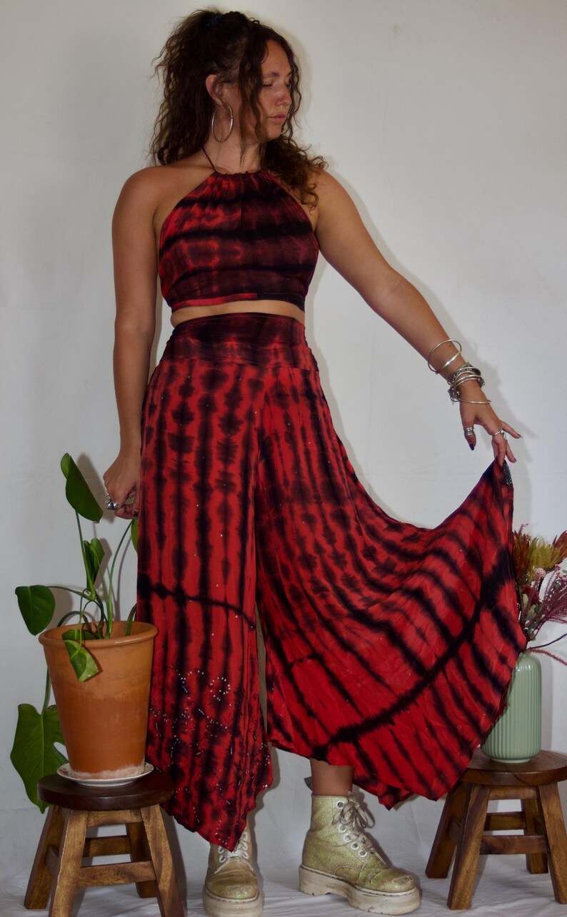 May include: A two-piece tie-dye outfit in red and black. The halter top and wide-leg pants feature a striped pattern. The pants have a beaded embellishment near the hem. The outfit is paired with light-colored boots.
