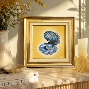 May include: A square painting in a gold frame featuring a blue iridescent seashell holding a single white pearl against a solid yellow background. The artwork is displayed on a surface with books, a ceramic pitcher, and a glass candle holder.