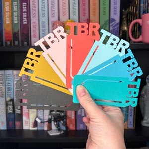 Puede incluir: Una mano sostiene un abanico de marcapáginas de papel de colores, cada uno con las letras "TBR" recortadas. Los marcapáginas son en tonos de rosa, amarillo, rojo, azul y turquesa. Estanterías llenas de libros están en el fondo.