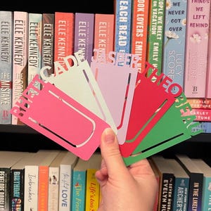 May include: Five colourful paper bookmarks with different names written on them. The bookmarks are in shades of pink, green, red, purple, and light blue.