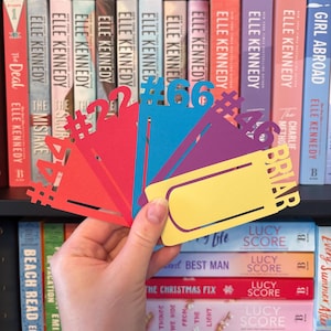 Op de afbeelding: Een hand houdt een waaier van kleurrijke papieren bladwijzers in rood, oranje, blauw, paars en geel vast. De bladwijzers hebben nummers en de naam "BRIAR" erop. Boekenplanken zijn op de achtergrond te zien.