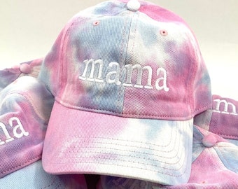 May include: A stack of pink and blue tie-dye baseball caps with the word "mama" embroidered in white on the front of the top cap.