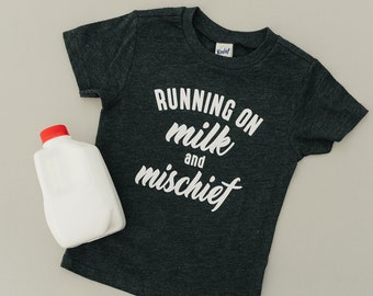 May include: A dark gray t-shirt with the text "RUNNING ON milk and mischief" in white. A white milk carton with a red lid is next to the shirt.