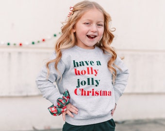 May include: A young child wearing a gray sweatshirt with the words "have a holly jolly Christmas" in green and red. The child is also wearing dark green pants and a plaid scrunchie. The child is smiling.