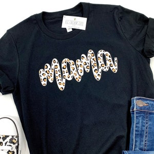 May include: A black t-shirt with a white 'mama' graphic design. The graphic design is in a leopard print pattern with brown and white spots.