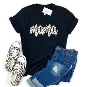 May include: Black t-shirt with a white "mama" graphic in a leopard print design. The shirt is paired with blue jeans and leopard print sneakers.