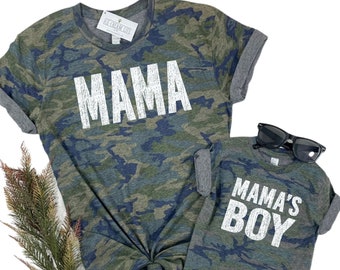 May include: Matching camouflage print t-shirts for a parent and child. The adult shirt says "MAMA" in white text and the child's shirt says "MAMA'S BOY" in white text.