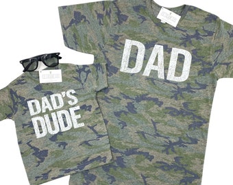 May include: Two camouflage t-shirts with white text. The larger shirt says "DAD" and the smaller shirt says "DAD'S DUDE".
