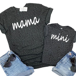 May include: A matching set of two t-shirts with a black leopard print. The larger shirt says "mama" in white script and the smaller shirt says "mini" in white script.